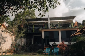 Edifício de dois andares com varanda, janelas amplas e área de jardim com cadeiras.
