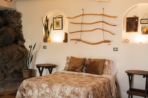 Quarto com cama, duas mesas de cabeceira, parede de pedra e prateleiras fixas decoradas com plantas e quadros.