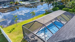 A modern outdoor swimming pool with a glass enclosure, surrounded by a well-maintained lawn and a body of water with lily pads. - Brand-New Cape Coral Oasis 4BR/3BA + Heated Saltwater Pool + Waterfront (Cape Coral)