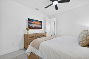A bedroom with a bed, a dresser, a ceiling fan, and a painting of a sunset over the ocean. - Brand-New Cape Coral Oasis 4BR/3BA + Heated Saltwater Pool + Waterfront (Cape Coral)
