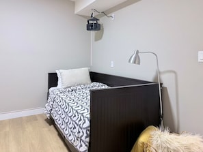 Uma cama de solteiro com um cobre-leito estampado, uma luminária de cabeceira e um projetor fixado no teto.