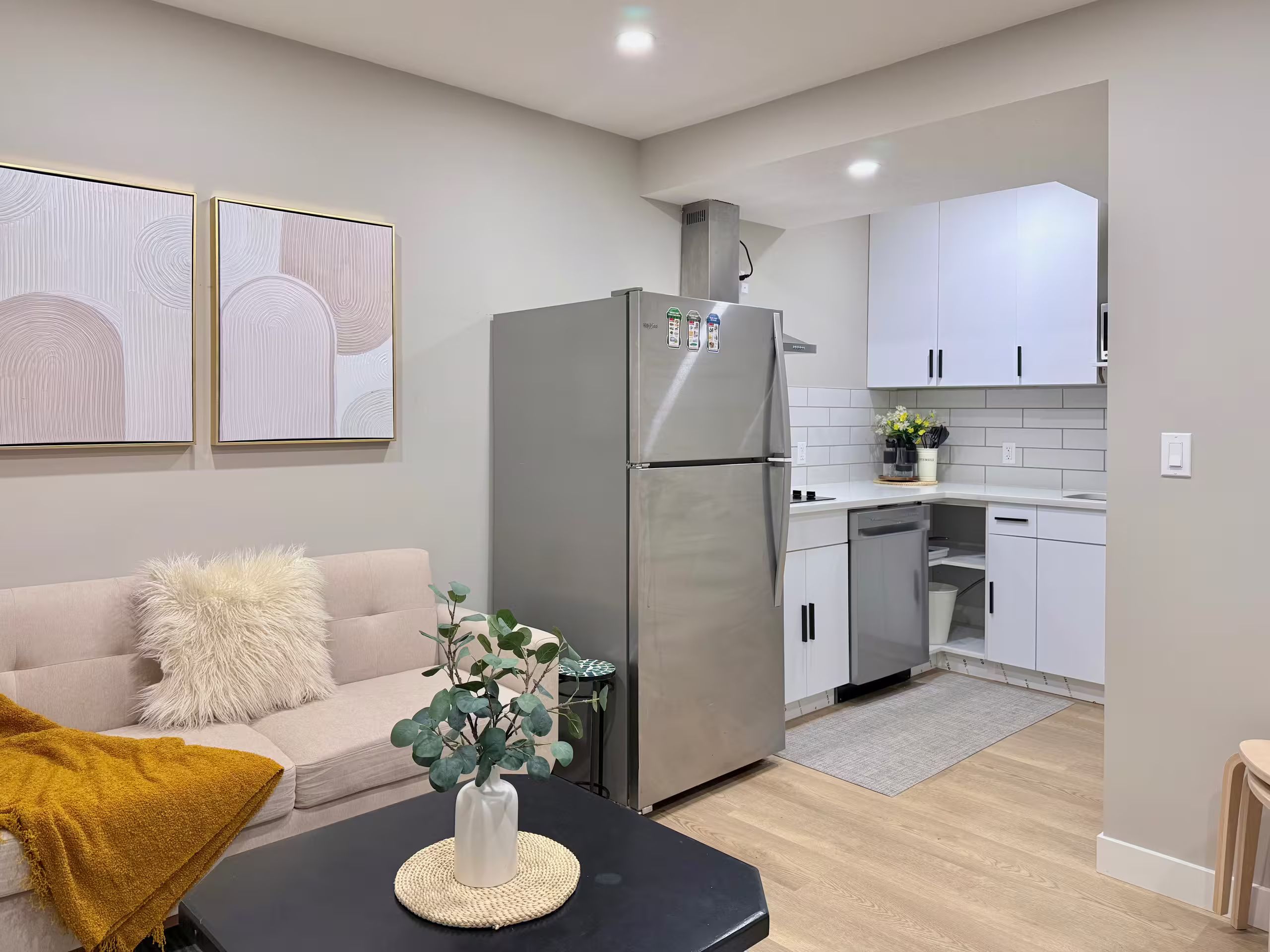Une cuisine moderne avec des meubles blancs, un réfrigérateur en acier inoxydable et un coin repas comprenant une table noire et un canapé recouvert d’un plaid jaune.