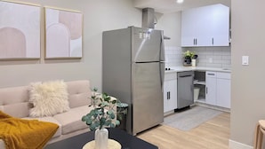 Une cuisine moderne avec des meubles blancs, un réfrigérateur en acier inoxydable et un coin repas comprenant une table noire et un canapé recouvert d’un plaid jaune.