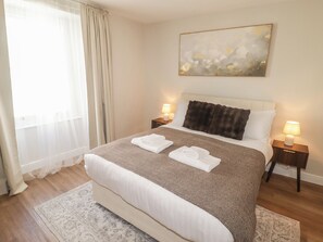 A hotel room with a large bed, two bedside tables with lamps, a window with sheer curtains, and a painting on the wall. - Flat 3 (Tenby)