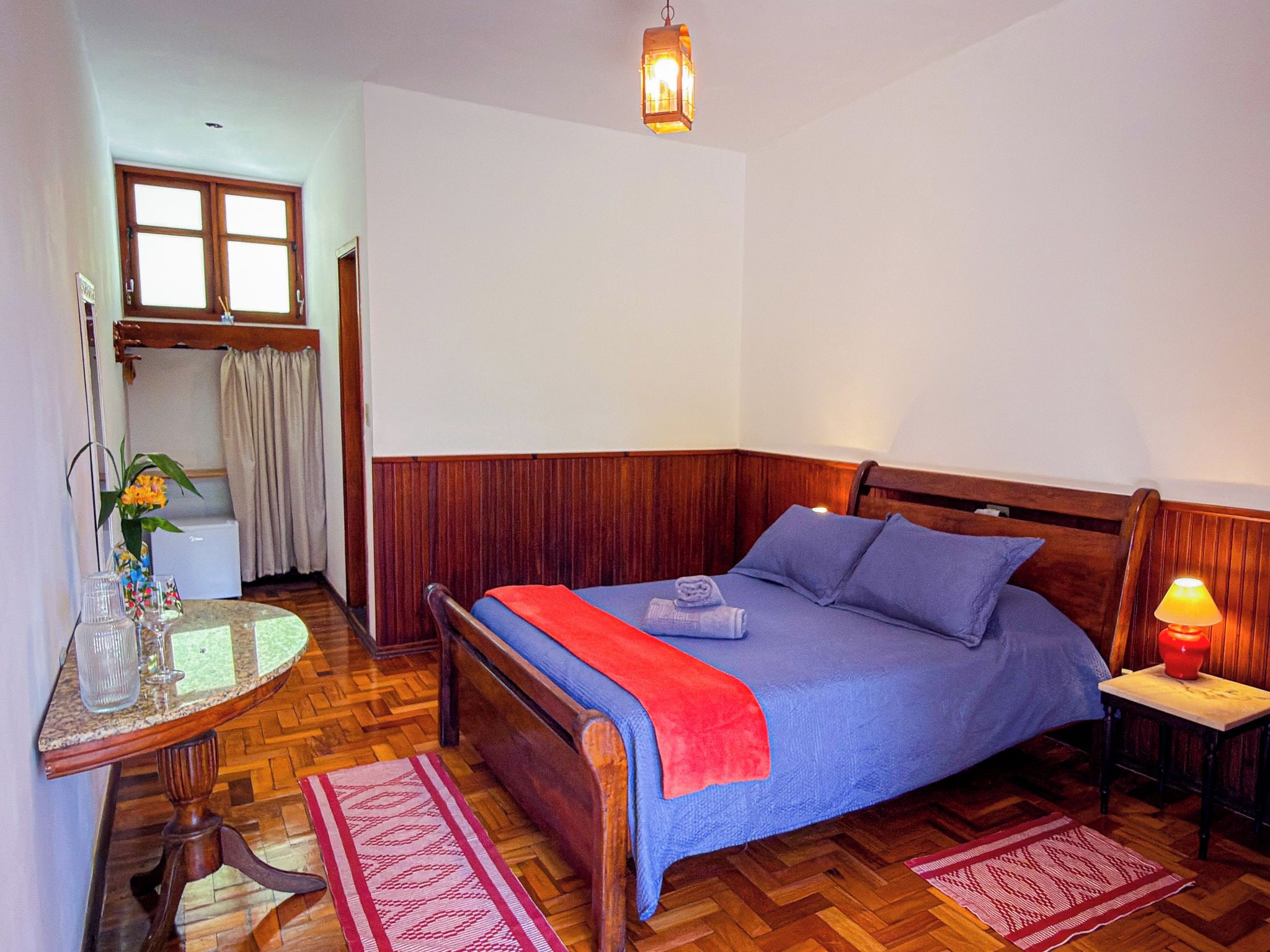 A room with a bed, a table with a vase and glasses, a lamp, and a window with curtains.
