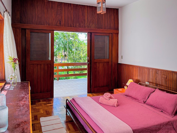 A bedroom with a bed, a nightstand, a window with wooden shutters, and a view of greenery outside.