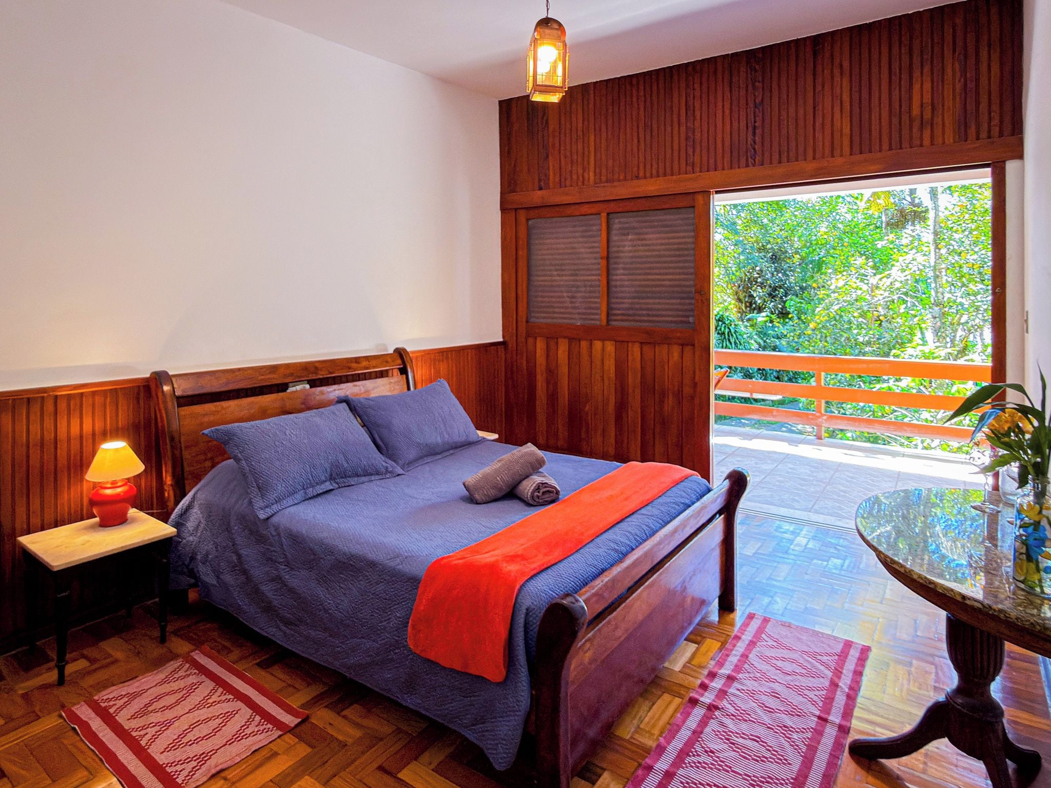 A bedroom with a wooden bed, a nightstand with a lamp, a window with a view of greenery, and a small table with a vase.