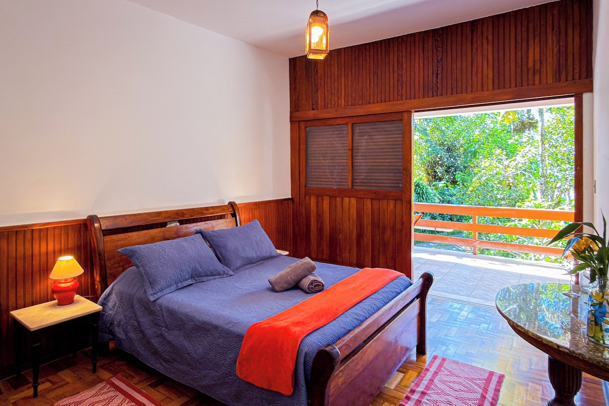 A bedroom with a wooden bed, a nightstand with a lamp, a window with a view of greenery, and a small table with a vase.