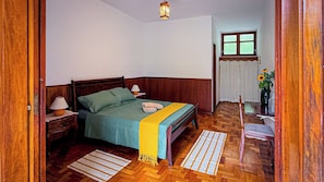 A bedroom with a wooden bed, a nightstand, a lamp, a window with curtains, and a wooden floor.