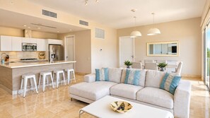 A modern living room with a sofa, a coffee table, and a kitchen area with a refrigerator, microwave, and bar stools. - Serene Retreat 2BR in Plaza Puntarena (Río Hato)