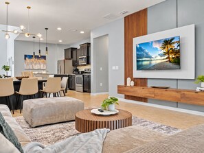 A modern living room with a dining area, kitchen, and a large TV mounted on the wall. - 6BR Disney Area Home | Pool, Spa & Resort (Kissimmee)