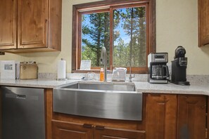 A kitchen with a stainless steel sink, wooden cabinets, a coffee maker, and a window with a view of trees. - Angel Fire Haus (Angel Fire)