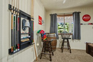 A room with a billiards rack, a billiard cue, a wooden barrel stool, and a window with curtains. - Angel Fire Haus (Angel Fire)