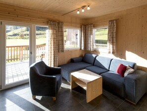 A room with a sectional sofa, a single armchair, and a small wooden table. There are two windows with curtains, and a deck is visible through the glass doors. - One-bedroom Cabin (Sankt Georgen am Kreischberg)