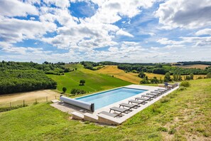 A swimming pool with lounge chairs, surrounded by green fields and trees under a partly cloudy sky.