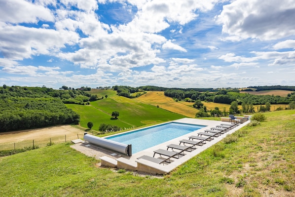 A swimming pool with lounge chairs, surrounded by green fields and trees under a partly cloudy sky.
