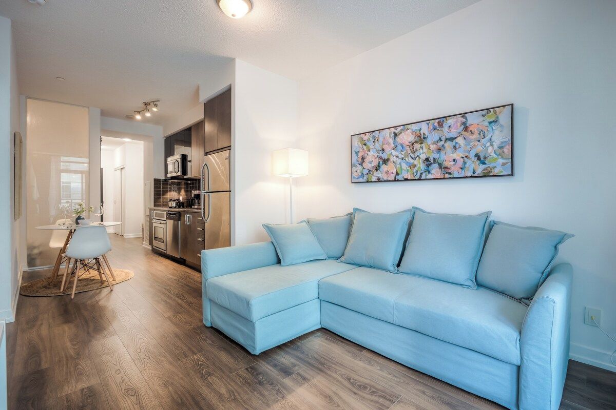 A modern living room with a light blue sectional sofa, a dining area with a white table and chairs, and a kitchen in the background.