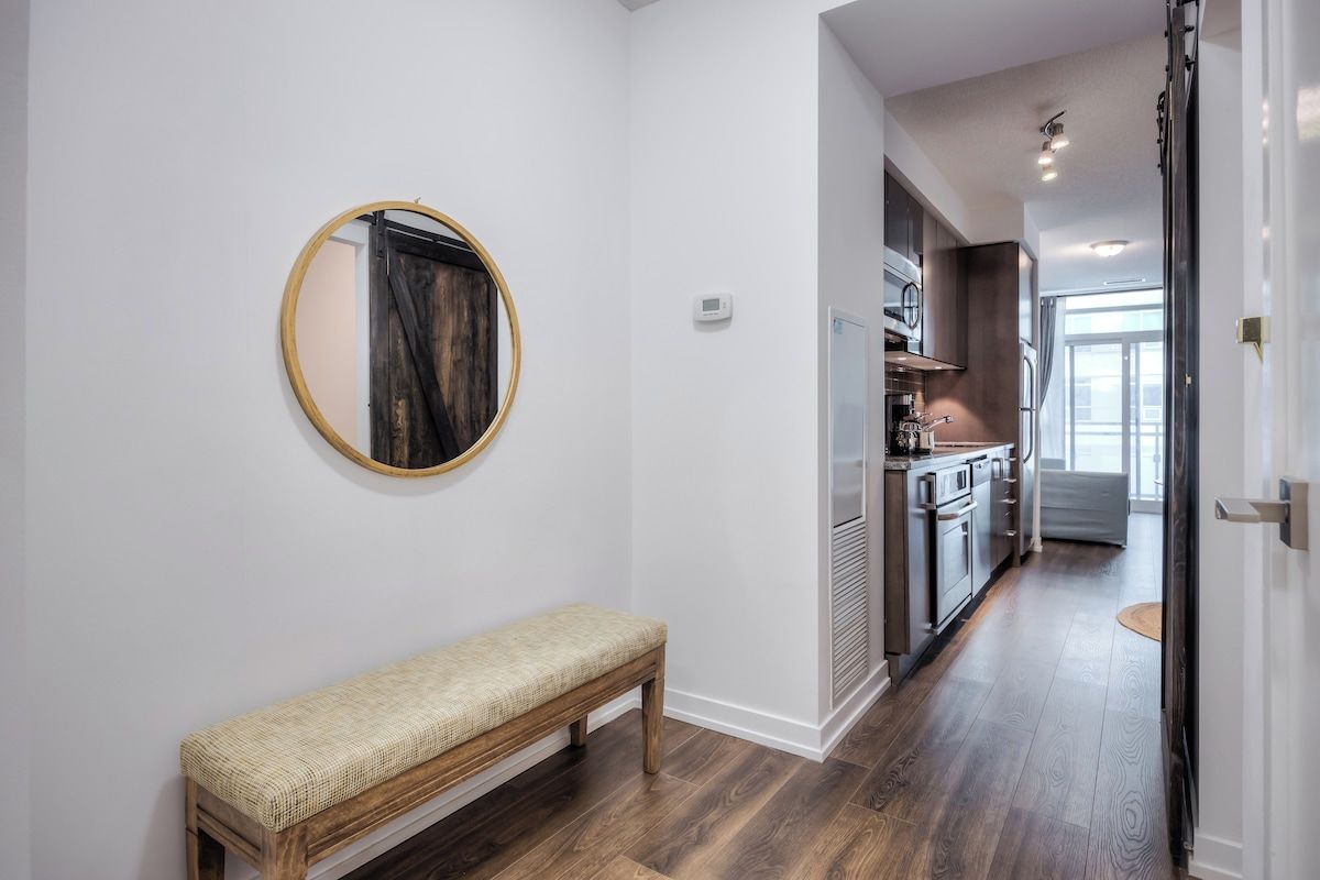 A hallway with a bench, a round mirror, and a kitchen area with cabinets and appliances.