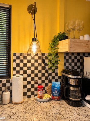 A kitchen with a checkered backsplash, a coffee maker, a bottle of coffee, a bag of coffee pods, a roll of paper towels, and a hanging light fixture.