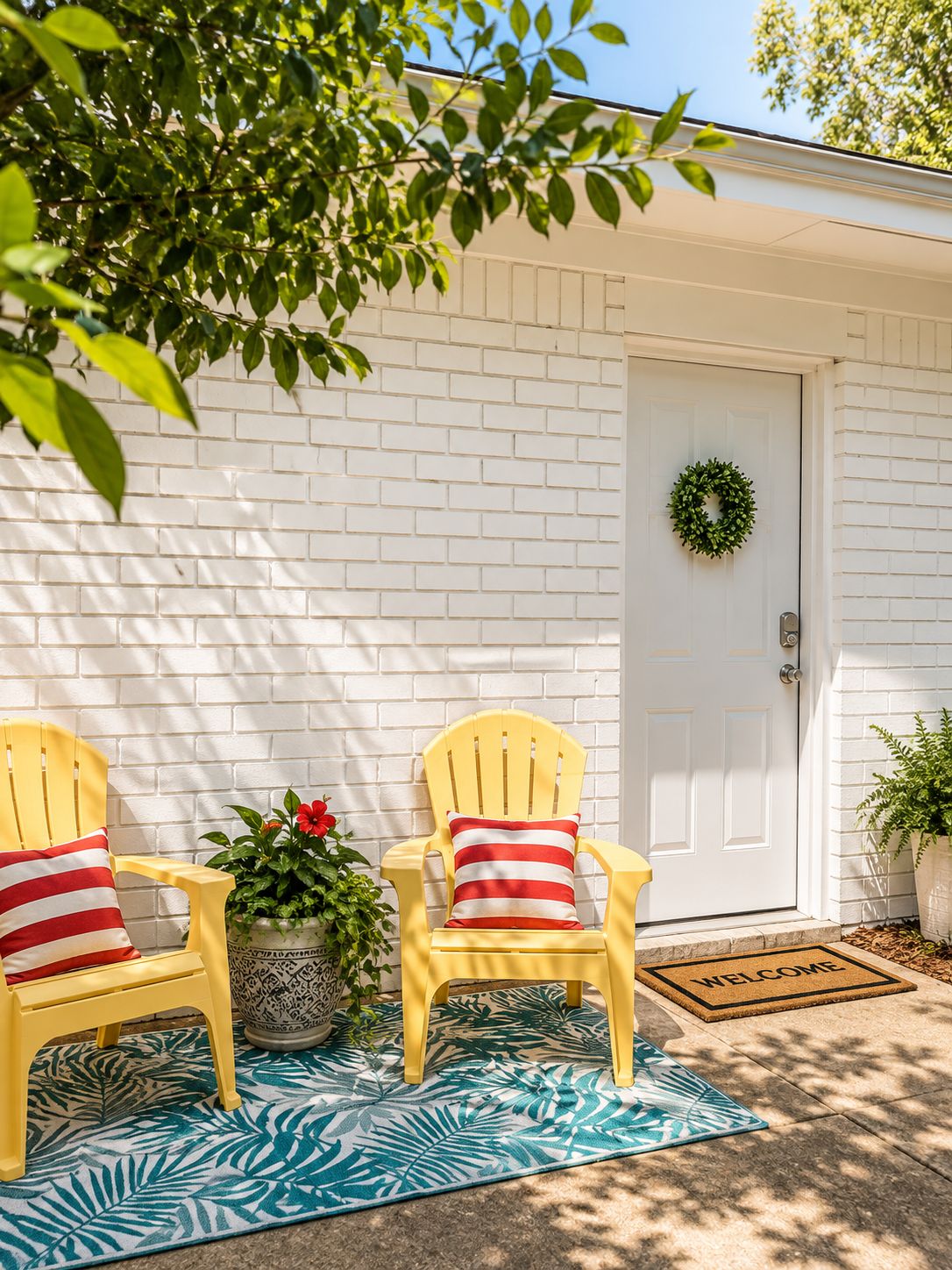 A white brick exterior with a white door, two yellow Adirondack chairs with striped pillows, a welcome mat, and a potted plant.