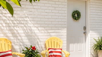 A white brick exterior with a white door, two yellow Adirondack chairs with striped pillows, a welcome mat, and a potted plant.