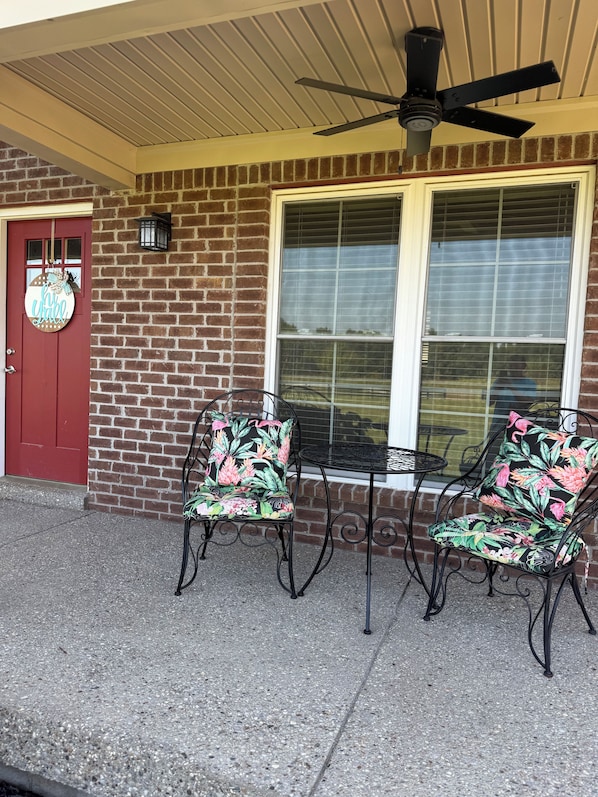A brick exterior with a red door, two outdoor chairs with floral upholstery, a ceiling fan, and a window with blinds.