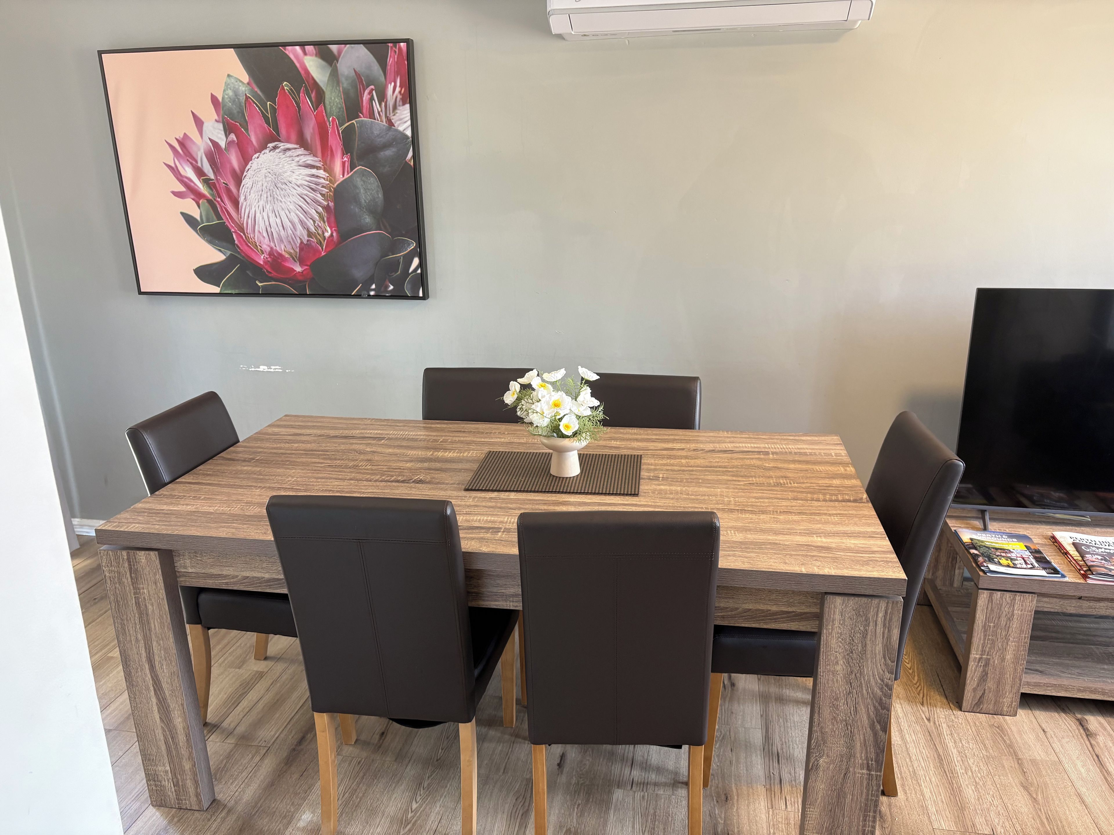 Un coin repas avec une table en bois, des chaises noires, un vase avec des fleurs et un téléviseur sur le côté droit.