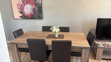 Un coin repas avec une table en bois, des chaises noires, un vase avec des fleurs et un téléviseur sur le côté droit.