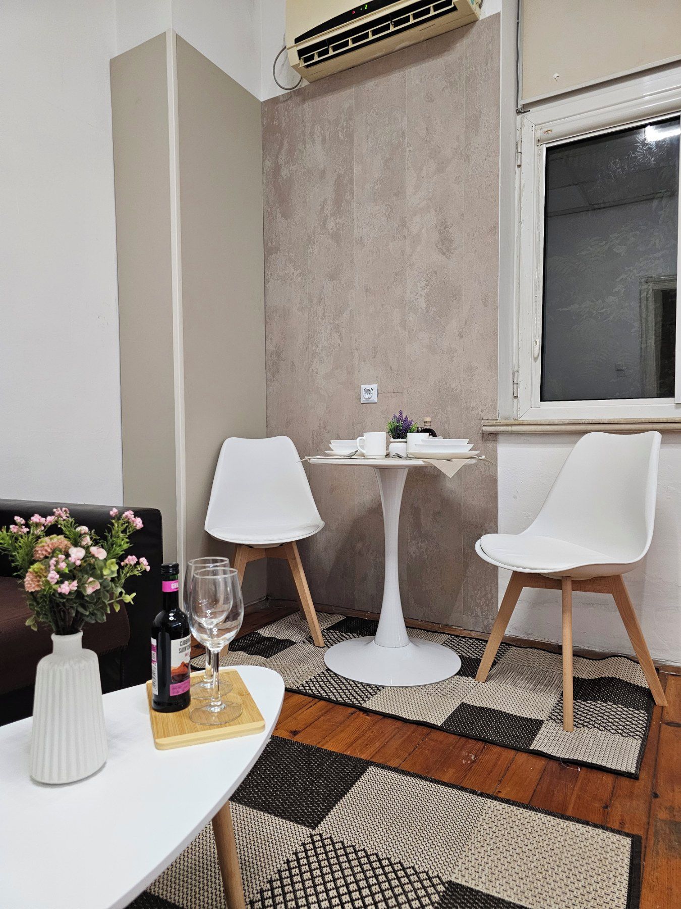 Un pequeño comedor con una mesa blanca, dos sillas blancas, una lámpara de mesa pequeña y blanca, un jarrón con flores y una botella de vino.