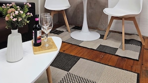 Un pequeño comedor con una mesa blanca, dos sillas blancas, una lámpara de mesa pequeña y blanca, un jarrón con flores y una botella de vino.