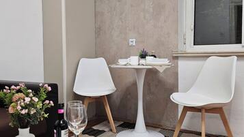 Un pequeño comedor con una mesa blanca, dos sillas blancas, una lámpara de mesa pequeña y blanca, un jarrón con flores y una botella de vino.