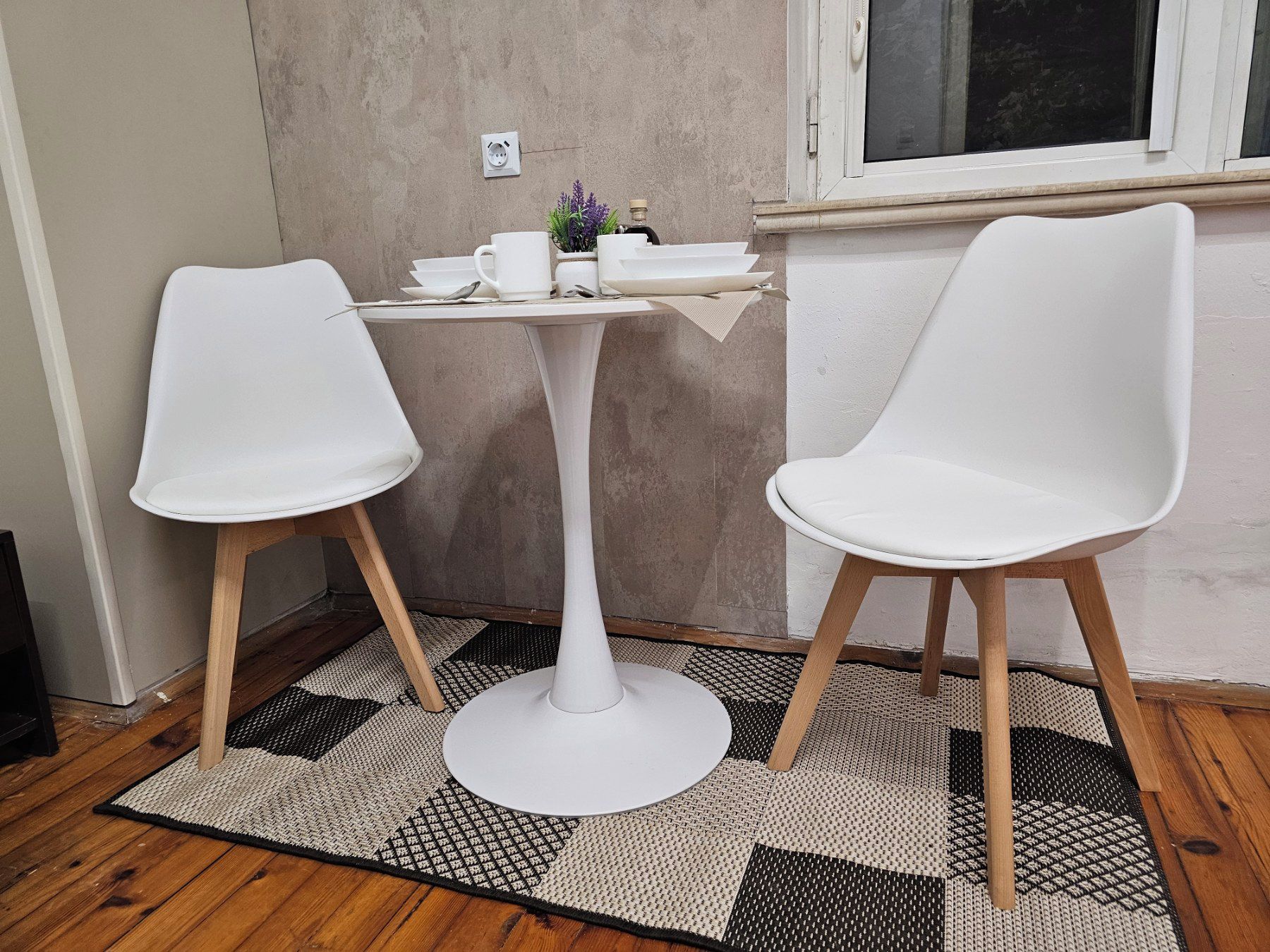 Una habitación pequeña con una mesa blanca y dos sillas blancas, una ventana y una alfombra con textura.