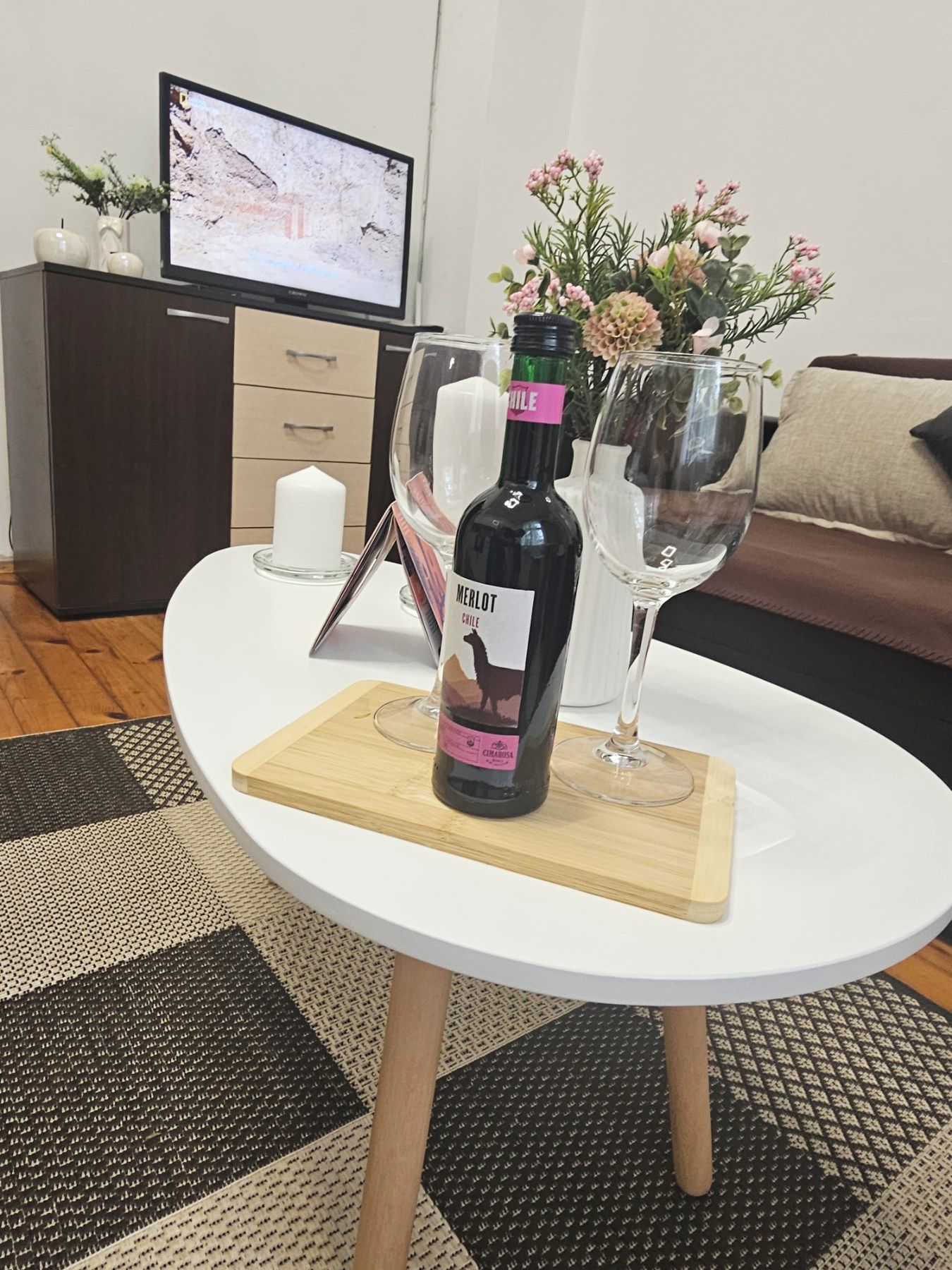 Uma garrafa de vinho Merlot sobre uma mesa branca com um apoio de madeira, uma taça de vinho e um vaso com flores.