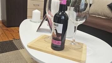 Uma garrafa de vinho Merlot sobre uma mesa branca com um apoio de madeira, uma taça de vinho e um vaso com flores.