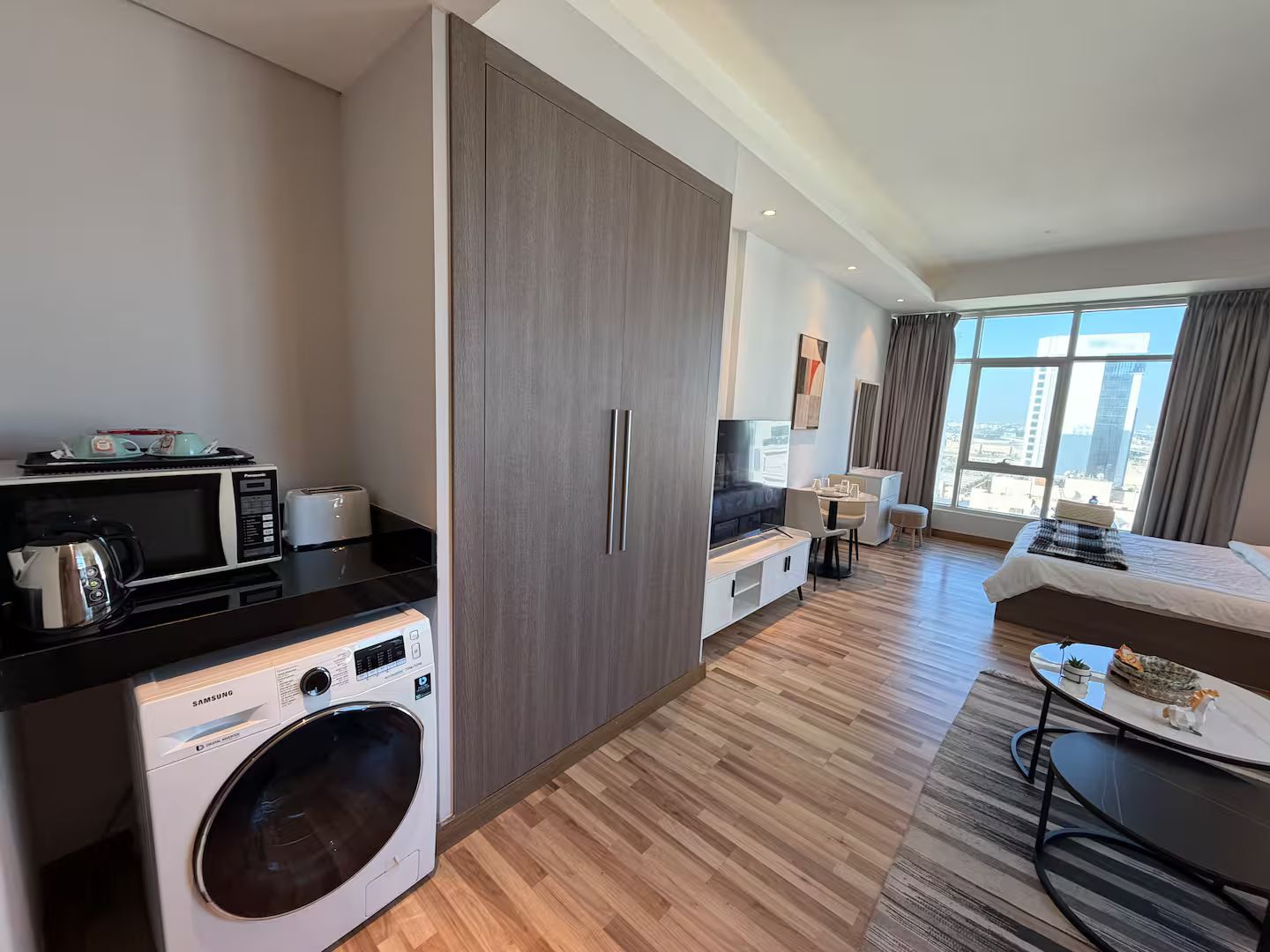 A modern hotel room with a large bed, a sofa, a dining area, a washing machine, and a view of the city.