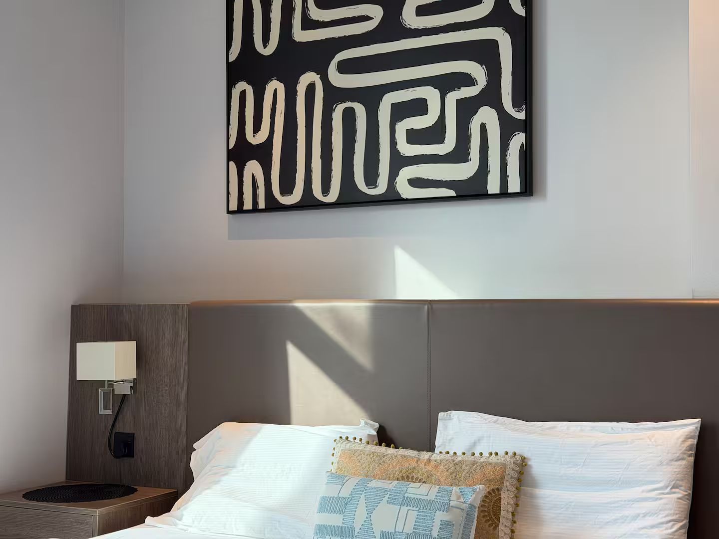 A modern hotel room with a large abstract artwork on the wall, a neatly made bed with two pillows, and a bedside table with a lamp.