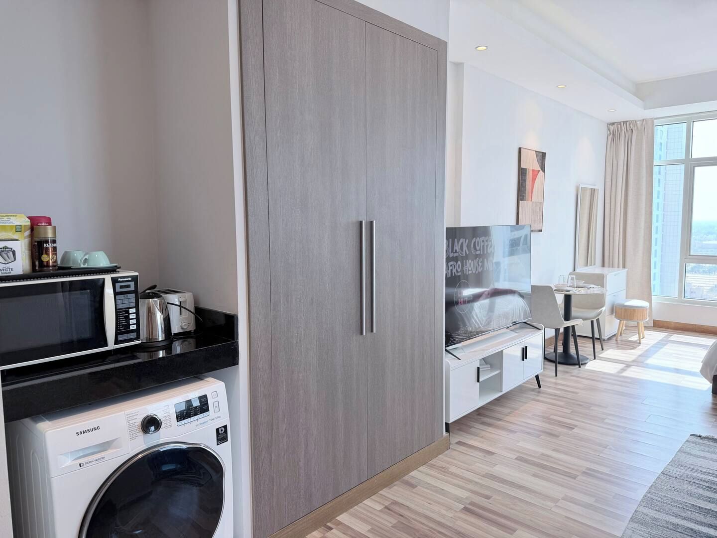 A modern hotel room with a washing machine, kitchenette, and a large wardrobe.