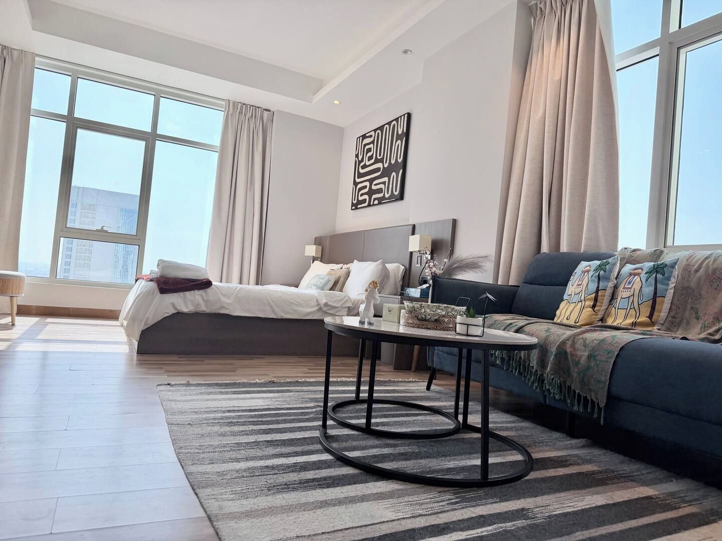 A modern hotel room with a large bed, a sofa, a coffee table, and a view of the city through large windows.