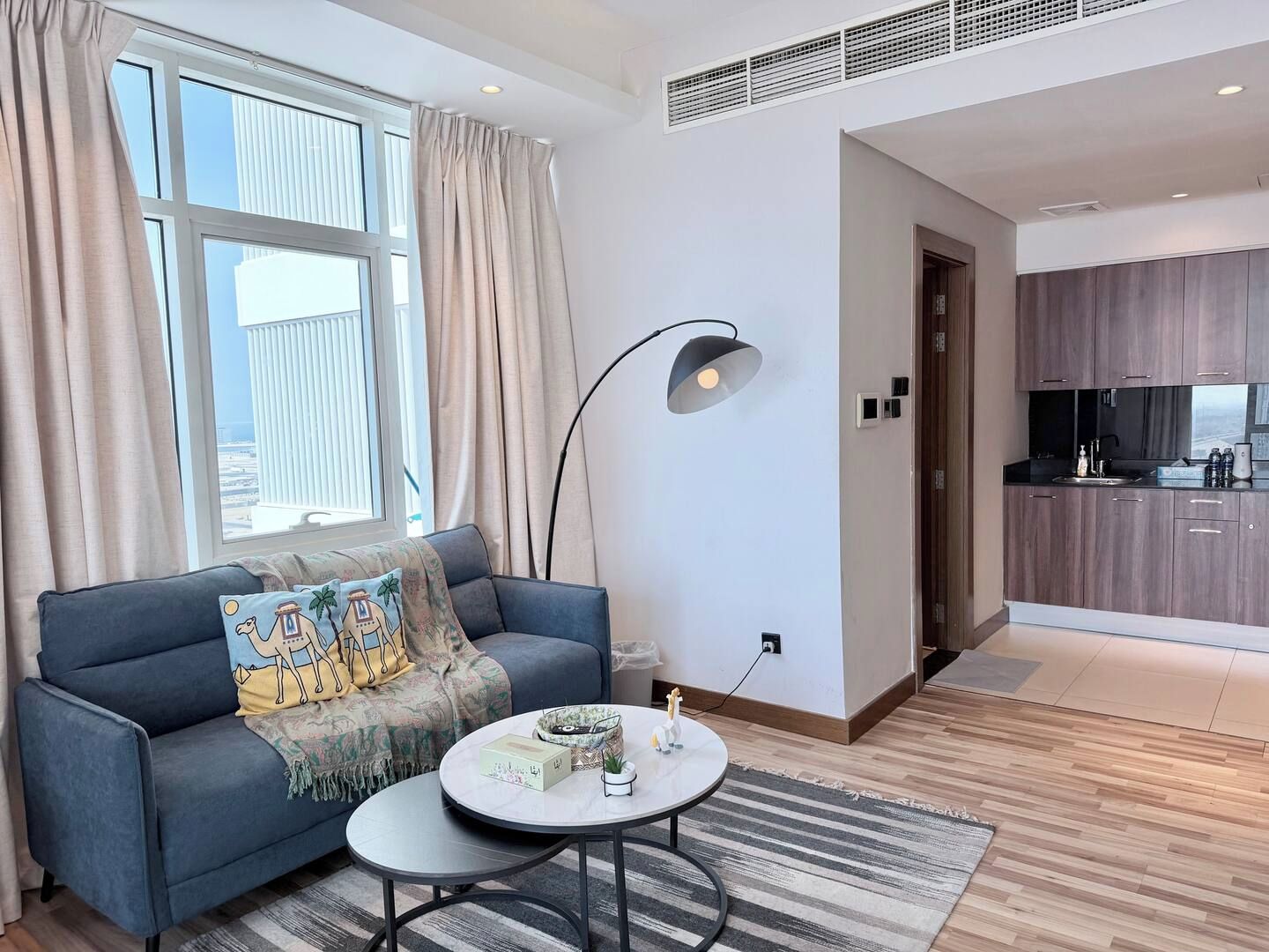 En moderne stue med sofa, sofabord og lampe. Et køkken er synligt gennem en åben dør.