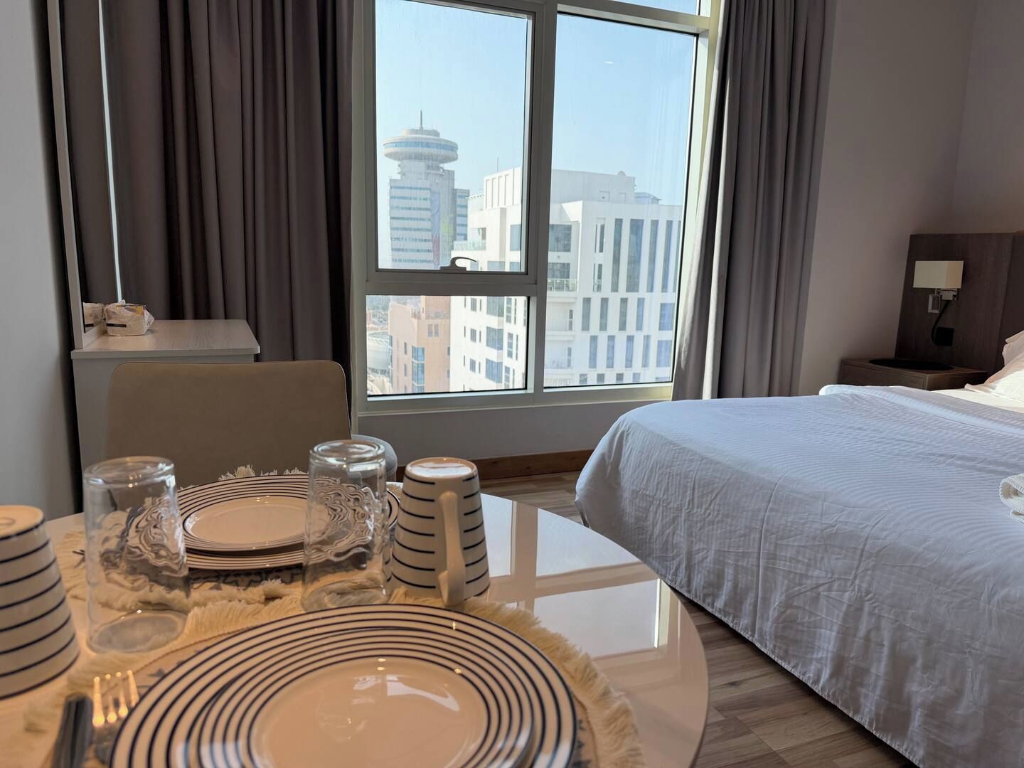 A hotel room with a bed, a dining table set with plates, glasses, and cutlery, and a view of buildings through the window.