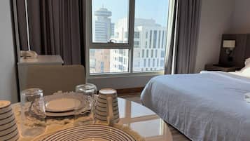 A hotel room with a bed, a dining table set with plates, glasses, and cutlery, and a view of buildings through the window.