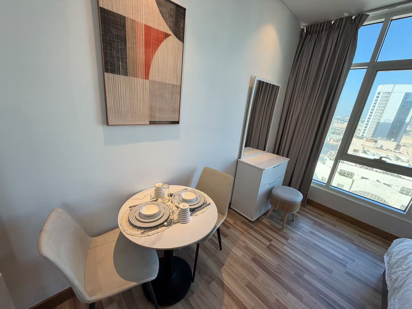 A hotel room with a round dining table set for two, a small white bedside table, a mirror, a window with curtains, and a city view.