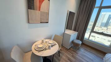 A hotel room with a round dining table set for two, a small white bedside table, a mirror, a window with curtains, and a city view.