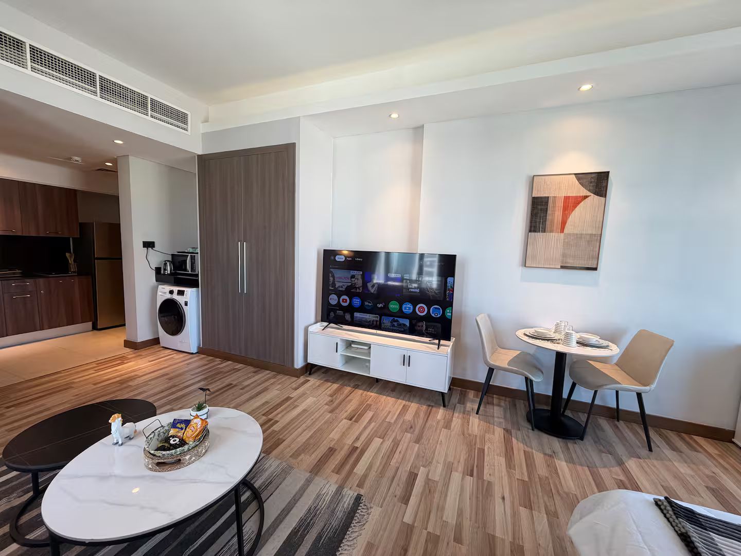 A modern living room with a flat-screen TV, a dining area with a table and chairs, a kitchen with a washing machine, and a large wardrobe.