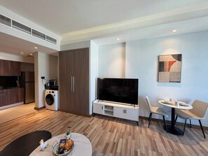 A modern living room with a flat-screen TV, a dining area with a round table and chairs, a kitchen with dark cabinets and a washing machine, and a ceiling with recessed lighting. - Best City View Studio / Heart of the city FNS1401O (Capital Governorate)