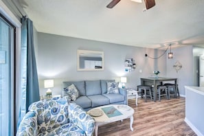 Ein Wohnzimmer mit grauem Sofa, einem gemusterten Sessel, einem kleinen runden Tisch und einem Essbereich mit Holztisch und Stühlen.