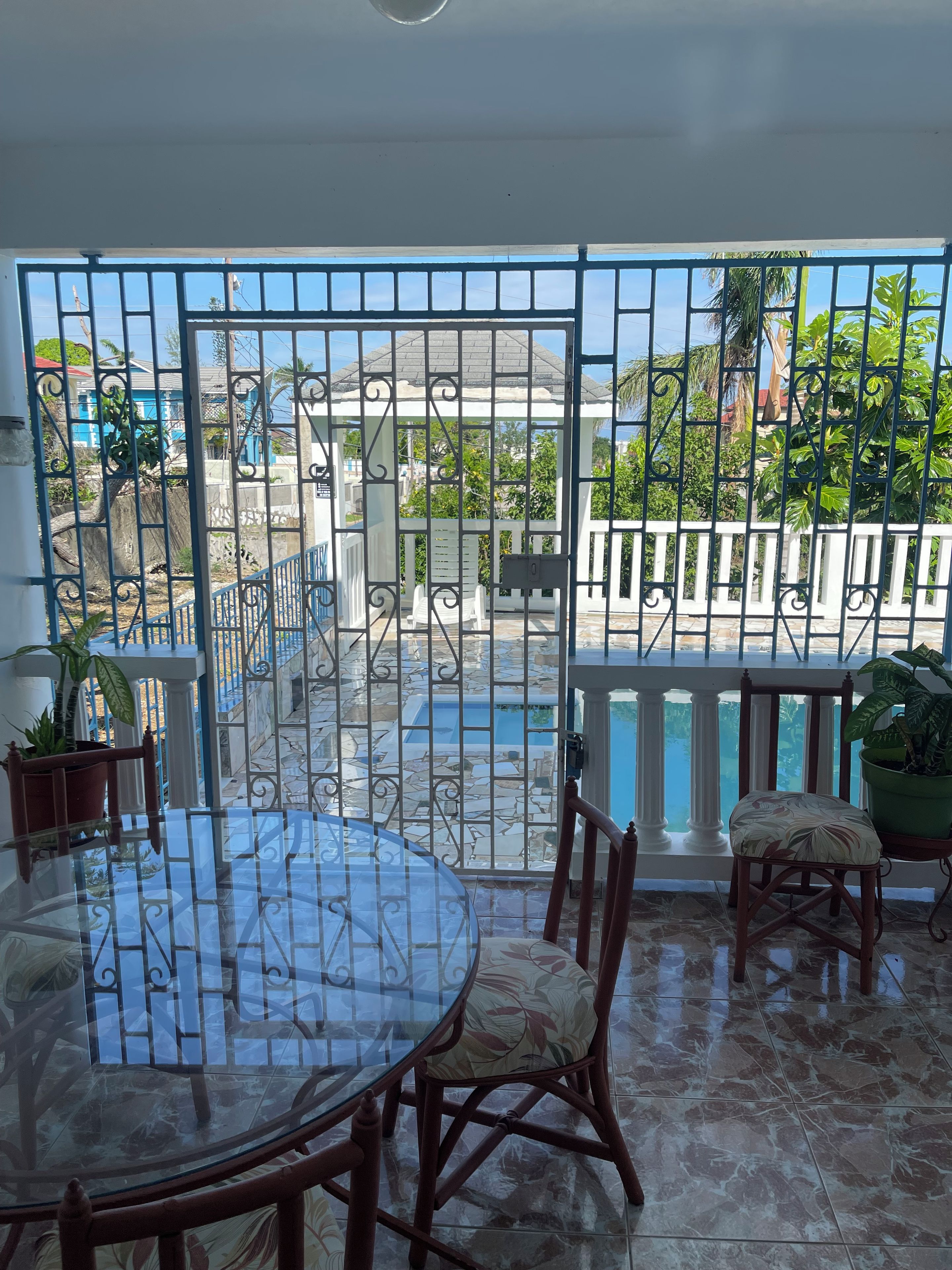 A balcony with a glass table and chairs, a view of a swimming pool, and a gated area with plants.
