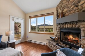 A living room with a stone fireplace, a large window with a view of mountains, and a door leading outside. - *New!* Big Sky Basecamp | Town Center & Hot Tub (Big Sky)