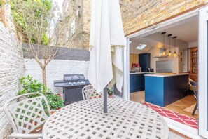 A patio with a round table and chairs, a grill, and a view into a modern kitchen with blue cabinets. - Cosy stylish 1 bed flat in Fulham w patio (London)