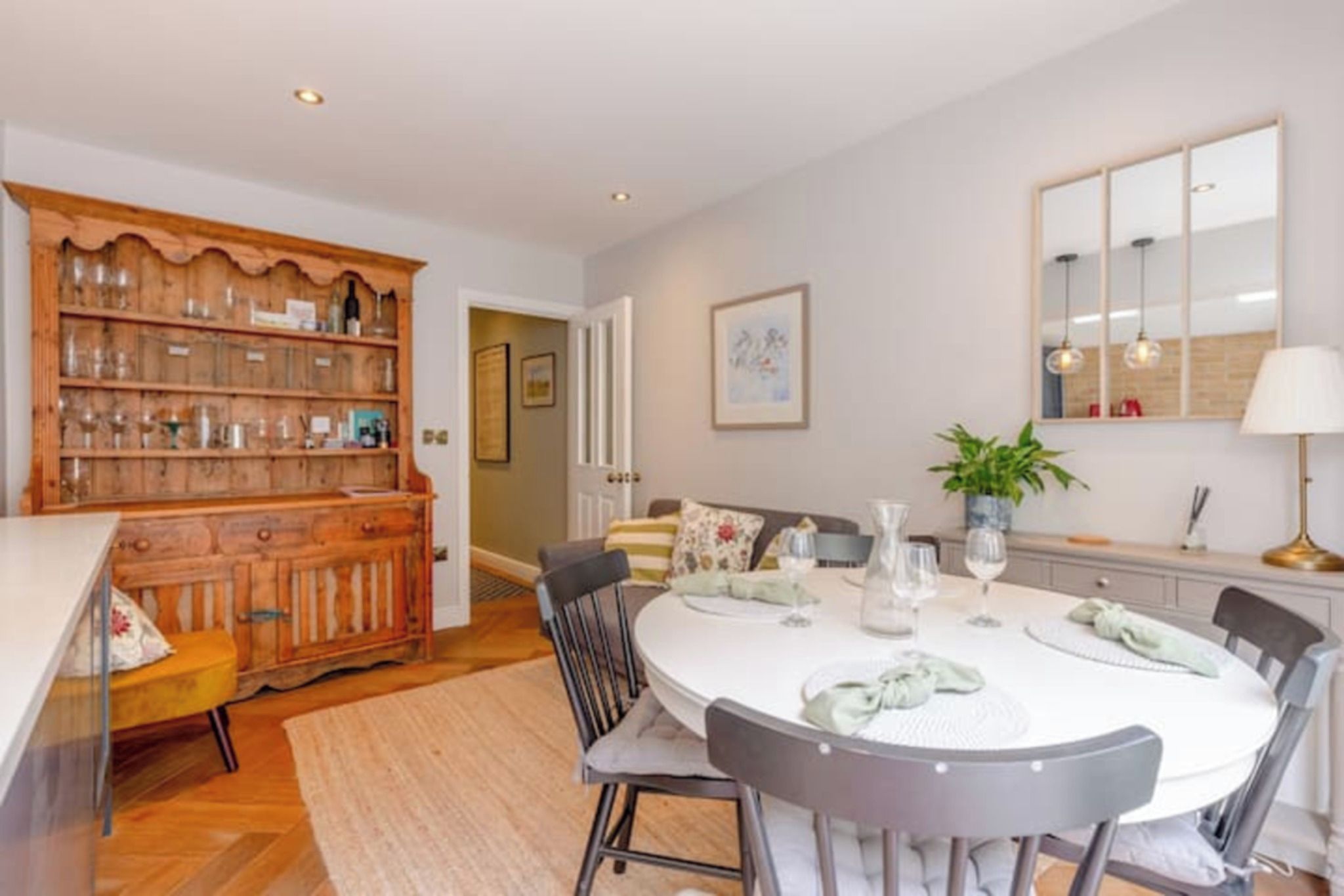 A dining area with a round table set for four, surrounded by chairs. A wooden cabinet with glassware is to the left.